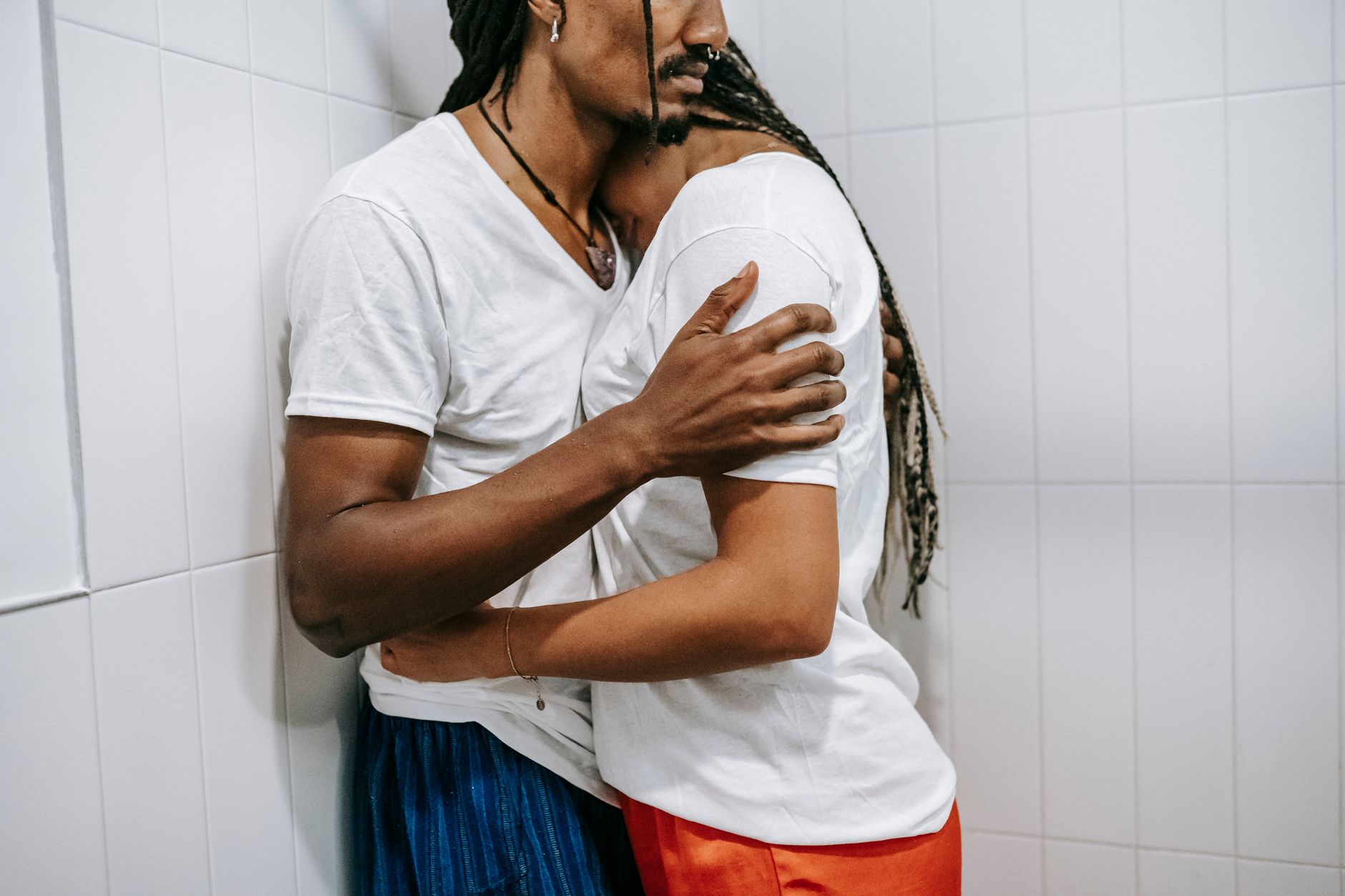 Side view of crop anonymous resentful young African American woman in casual clothes with dreadlocks hugging husband leaning on wall in bathroom
