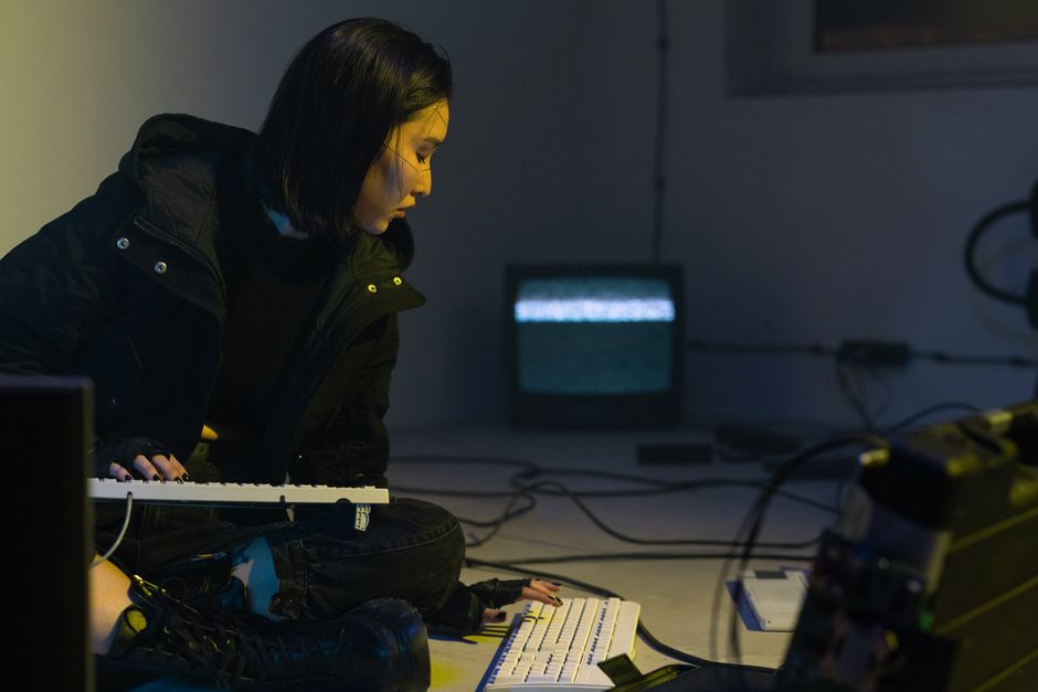 An Asian woman in a dark room works on a keyboard with a static-filled TV nearby.